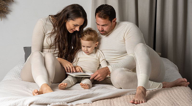 In the photo you can see the whole family – mom, dad, and a toddler – sitting on a bed, reading a book. They are all wearing natural color matching two-piece pajamas made from organic 100% Merino wool.