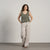 A smiling woman with long, curly brown hair wears a sleeveless linen crop top with side ties and light-colored wide-leg linen pants. She stands in front of a textured light gray wall, with one hand in her pocket.