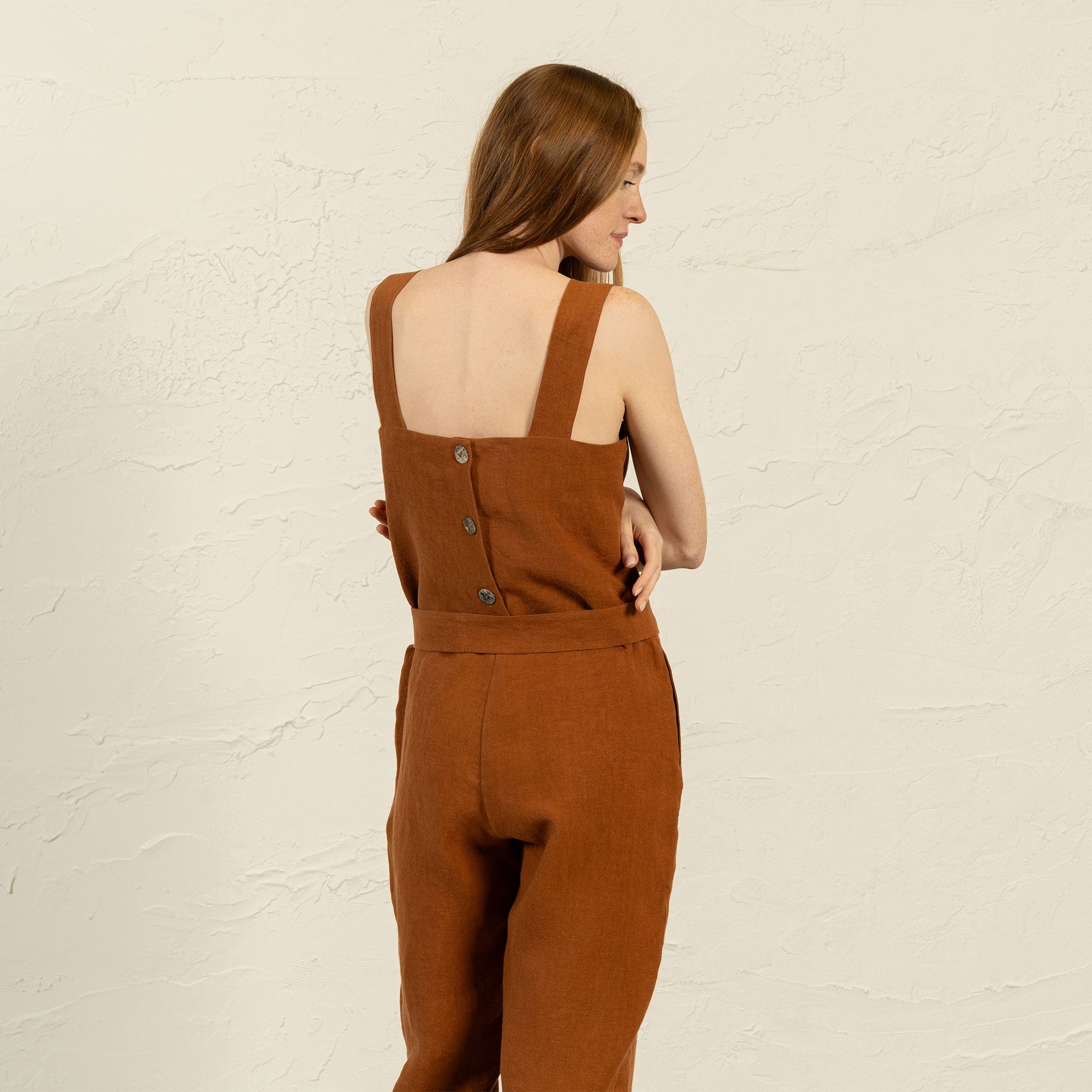 A light-skinned woman with long brown hair stands facing forward, wearing a linen menique jumpsuit with a tied belt at the waist and olive-green slip-on shoes. She has a neutral expression and her hands are resting on her hips.