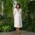 A smiling person with short, dark hair and what appears to be Asian features is holding a bouquet of light pink and white flowers. They are wearing a pure white wrap menique dress with elbow-length sleeves and are standing outdoors amidst lush green foliage and other flowers.