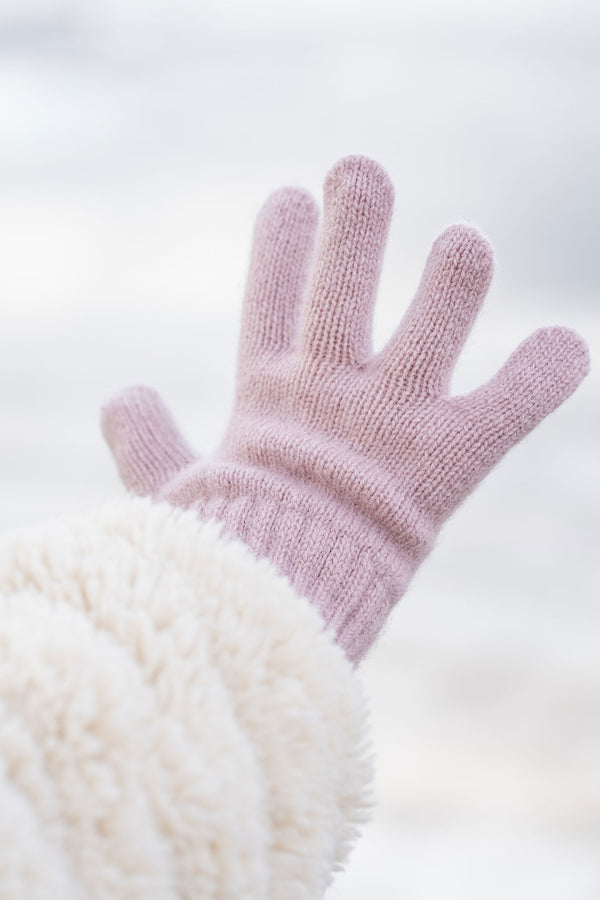A hand wearing a light pink knitted glove with a ribbed wrist extends upwards and slightly to the left, revealing a plush, cream-colored sleeve on the arm, against a blurred light background.